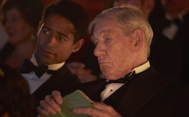 An elderly white man sit in a theater holding a green note pad.